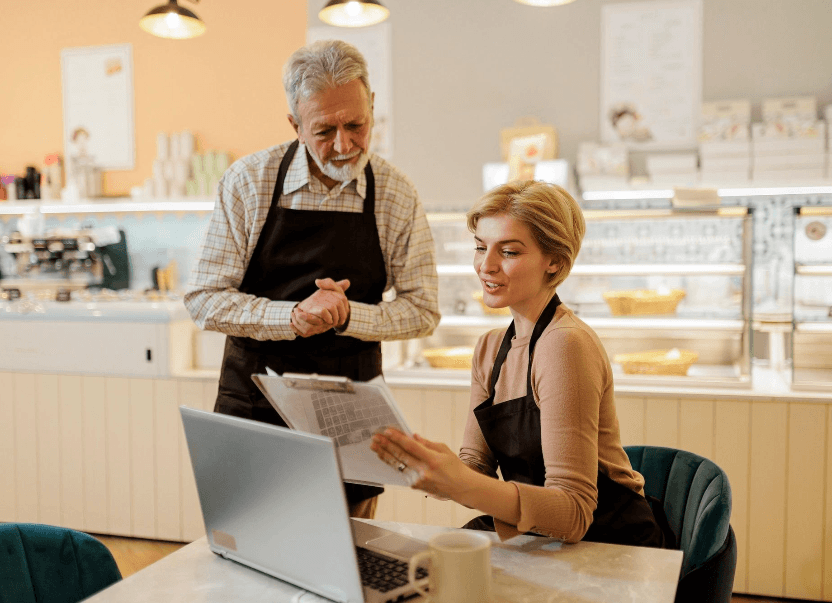 Two cafe staff in black aprons review documents on a clipboard and a laptop.